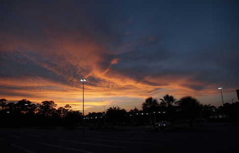 [Two photos stitched together with a huge cloud mass covering most of the sky. There is a open area of sky in the distance which lights the underside of this huge cloud as the sun sets. Sky color is blue/grey with beige/yellow patches. The lights above the nearly empty parking lot are lit putting white splotches on the image.]